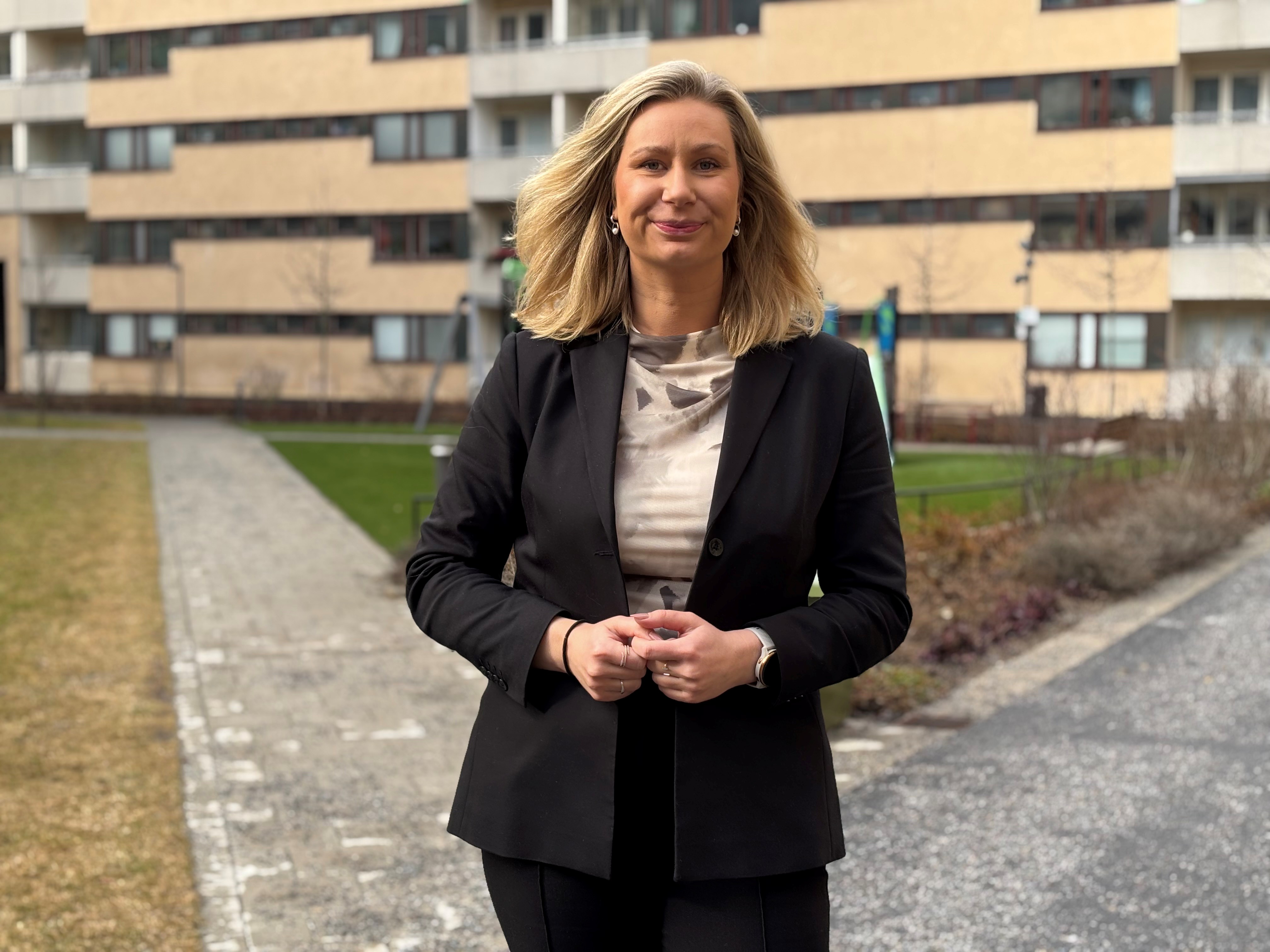 En blond kvinna i mörk kostym står på en innergård till ett flerbostadshus