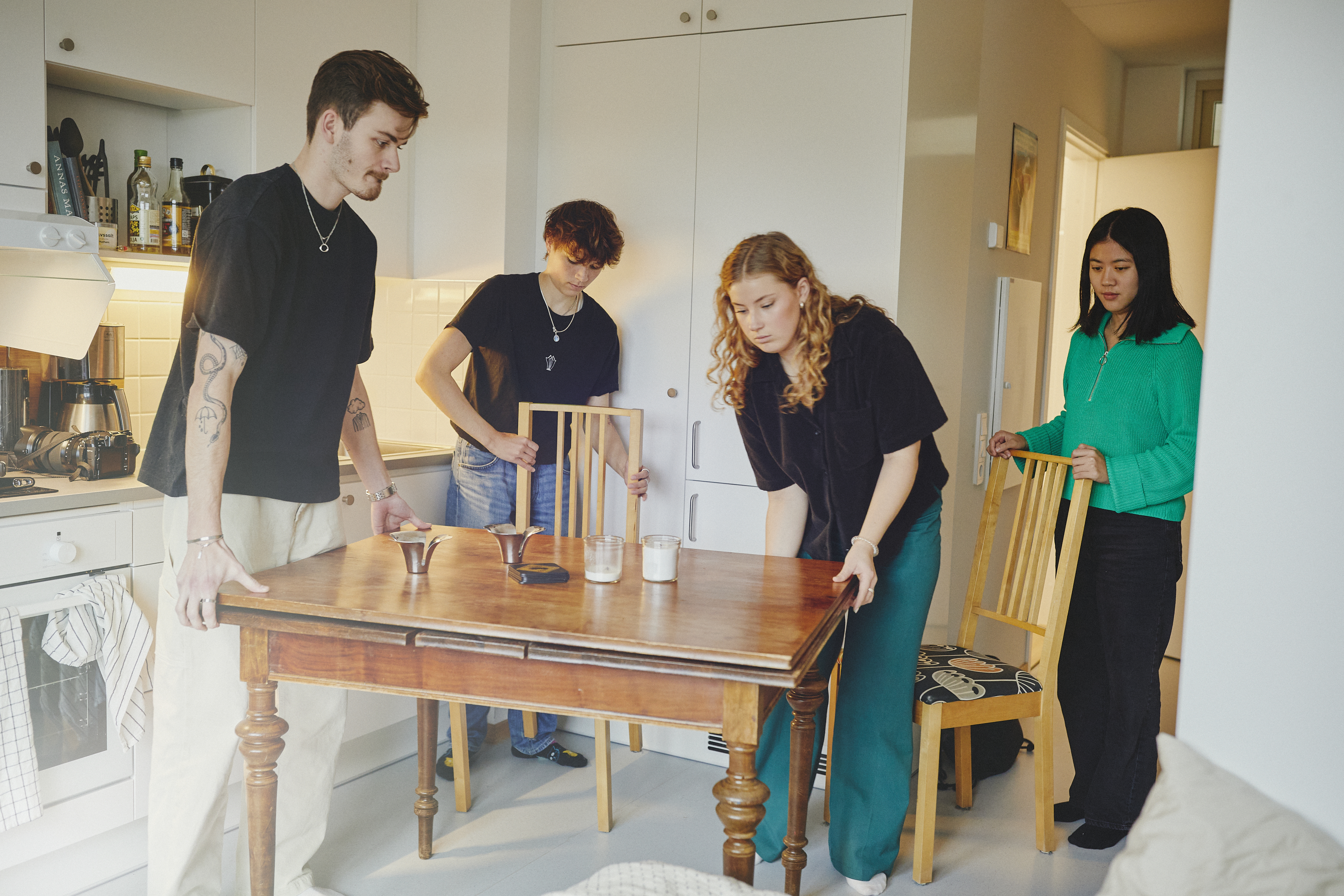 Fyra unga personer möblerar om i ett kök. De flyttar ett träbord och några stolar.