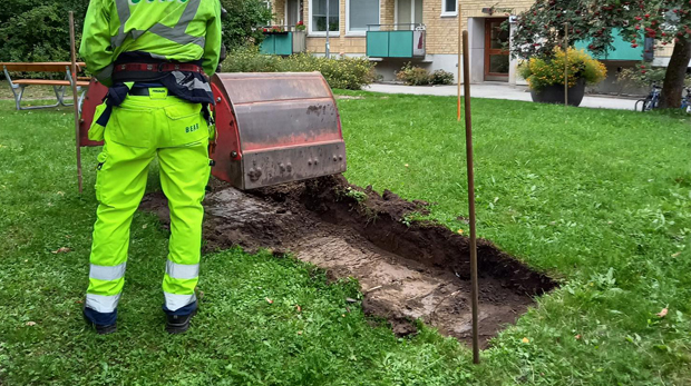 Man i varselkläder styr grävskopa som gräver upp del av gräsmatta