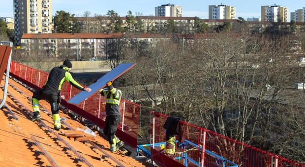 Två män arbetar på ett tak och lyfter upp en röd solpanel i luften.